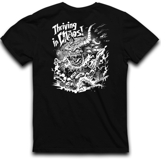 A CaliforniaScumbags Thriving In Chaos - Black Heavyweight Tee featuring a fierce, horned creature engulfed in flames, with buildings below being destroyed. The text above the creature reads, "Thriving in Chaos!" in a bold, dynamic font. The design is in white, creating a stark contrast against the shirt's boxy fit and black background.